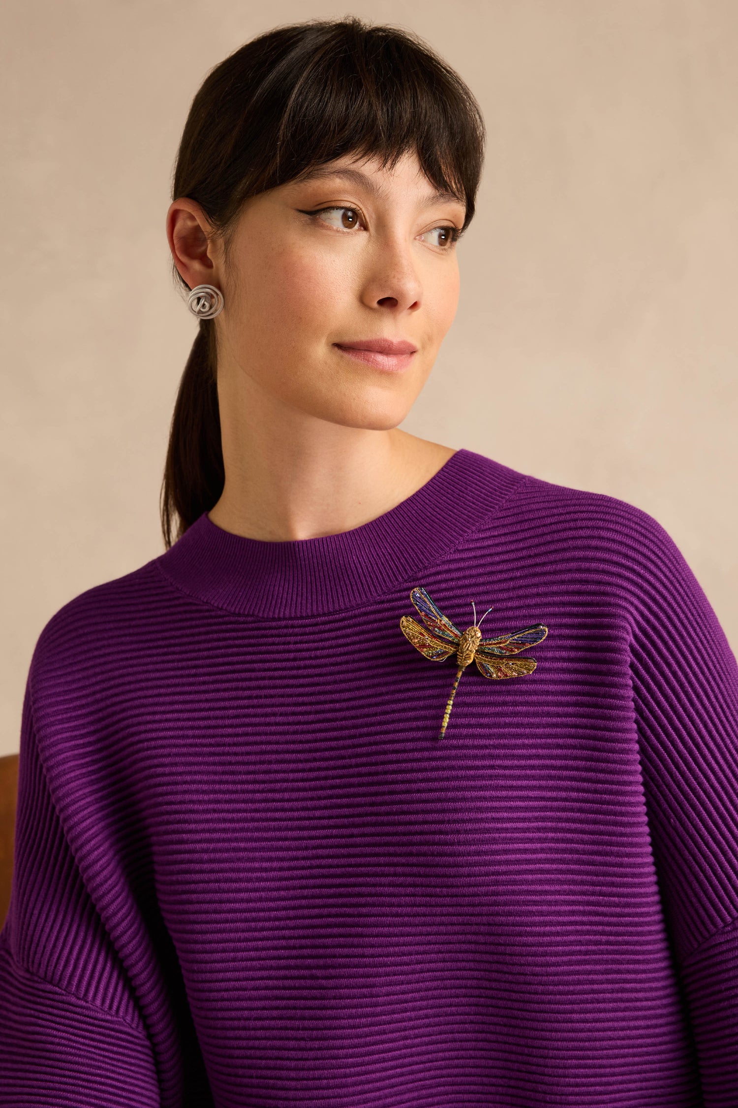 A woman wearing a ribbed purple sweater, styled with the Hand Beaded Hawker Dragonfly Brooch and silver circular earrings, gazes to the side against a neutral background.
