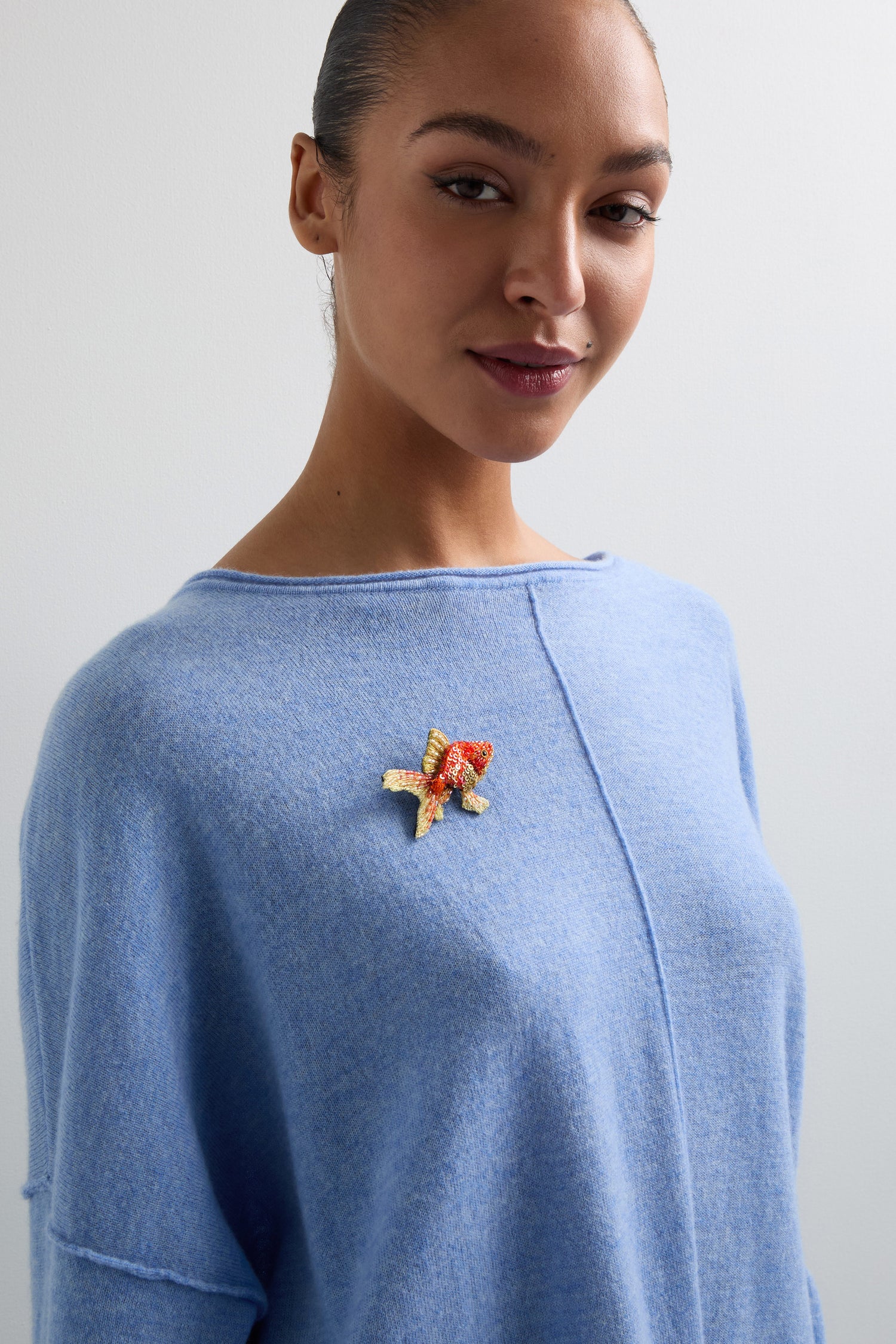 A person wearing a light blue sweater adorned with a Trovelore Hand Beaded Fantail Goldfish Brooch, standing against a plain background.