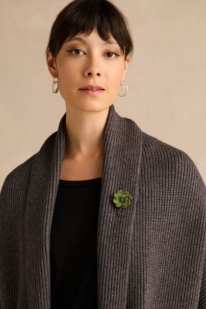 A woman with short brown hair, silver hoop earrings, a ribbed gray shawl, and a black top stands against a neutral background, her shawl adorned with the Hand Beaded Vintage Clover Brooch.