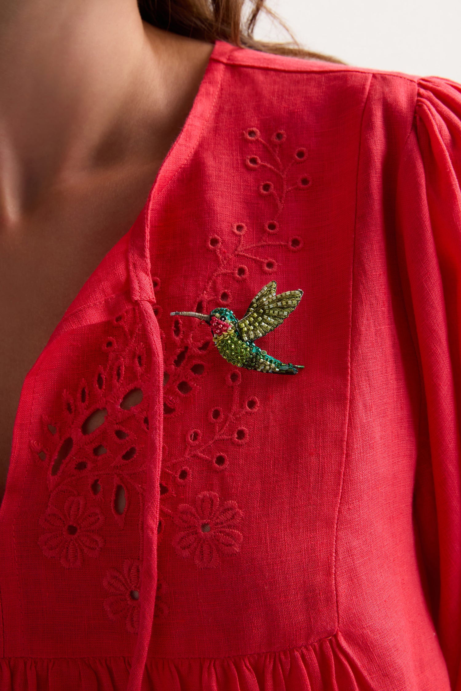A person wearing a bright red blouse with floral embroidery, featuring the Hand Beaded Ruby Throated Hummingbird Brooch pinned to the chest.
