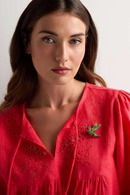 A woman stands against a plain white background, wearing a red embroidered blouse adorned with the Hand Beaded Ruby Throated Hummingbird Brooch pinned to her chest.