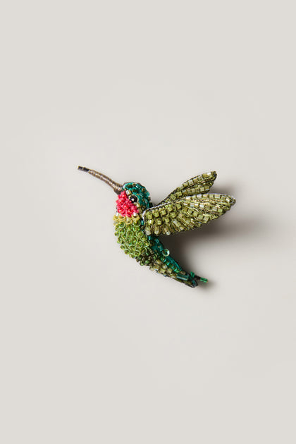 The Hand Beaded Ruby Throated Hummingbird Brooch features green, blue, and pink beads with metallic accents, handcrafted in a hummingbird design and set against a plain light background.