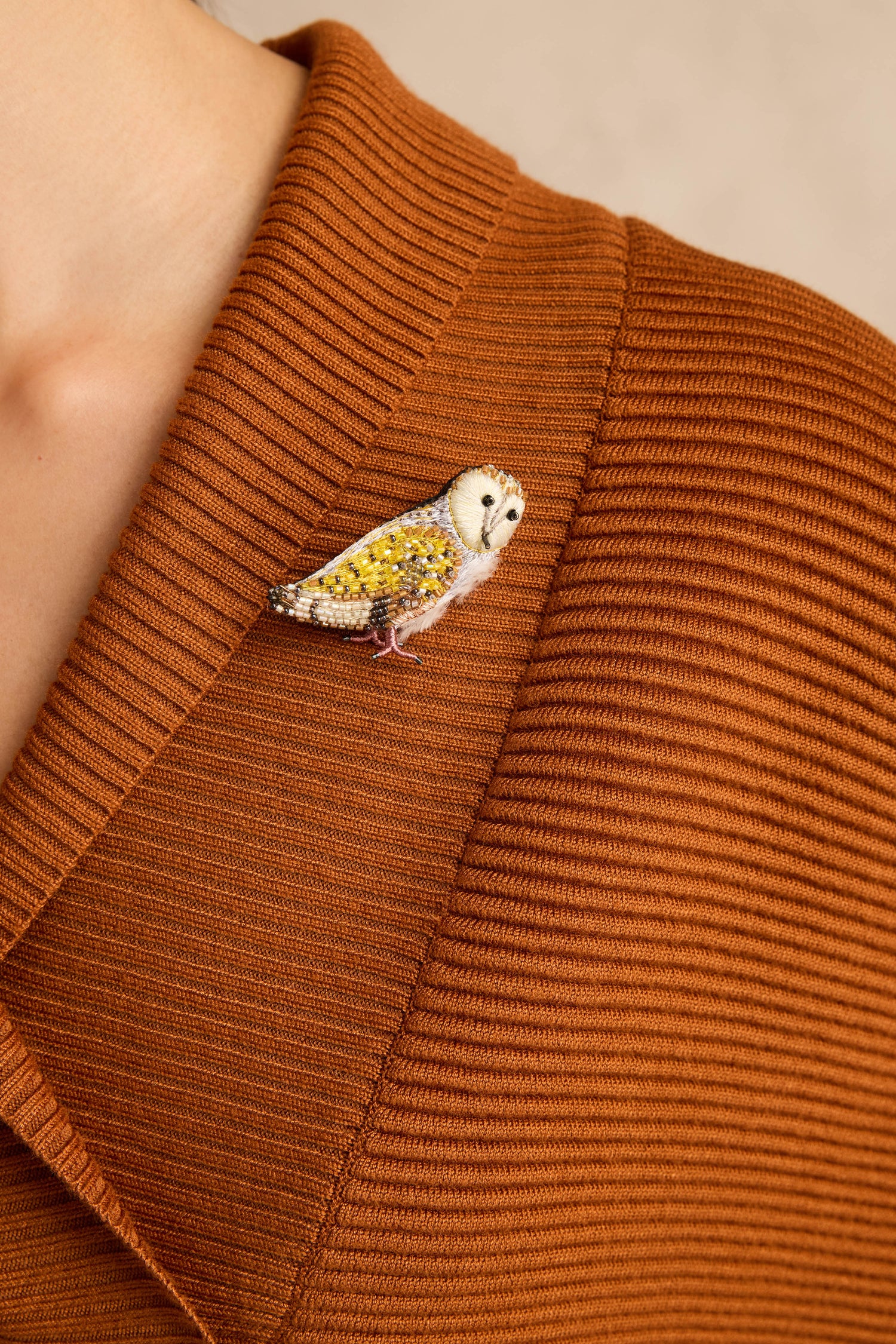 A close-up of a person wearing a ribbed, rust-colored garment with the Hand Beaded Gold Owl Brooch pinned to the collar.