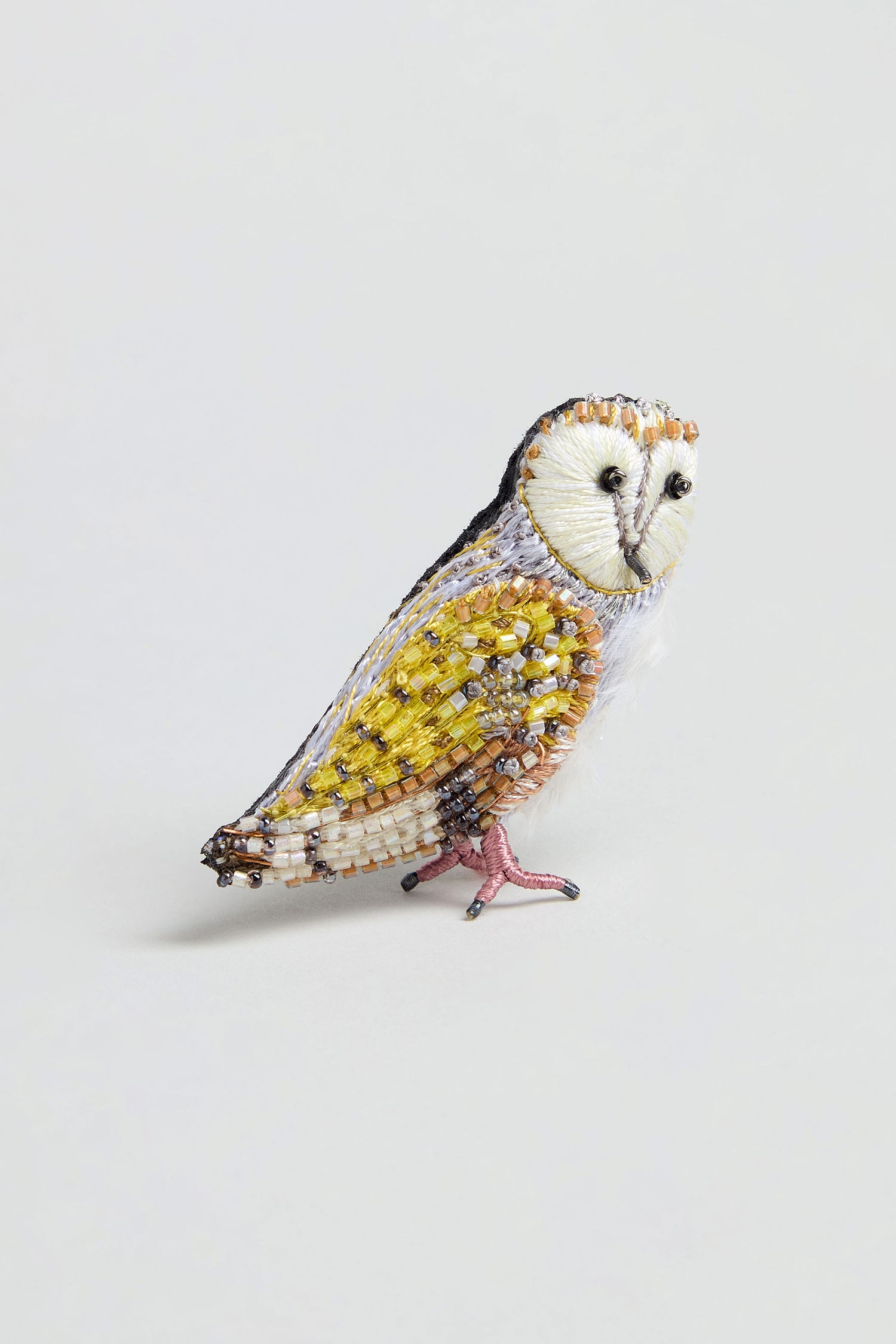 The Hand Beaded Gold Owl Brooch showcases intricate details with yellow, brown, white, and black beads in an owl design. Featuring a metal backing and set against a light background, this charming accessory adds a delightful accent to any outfit.