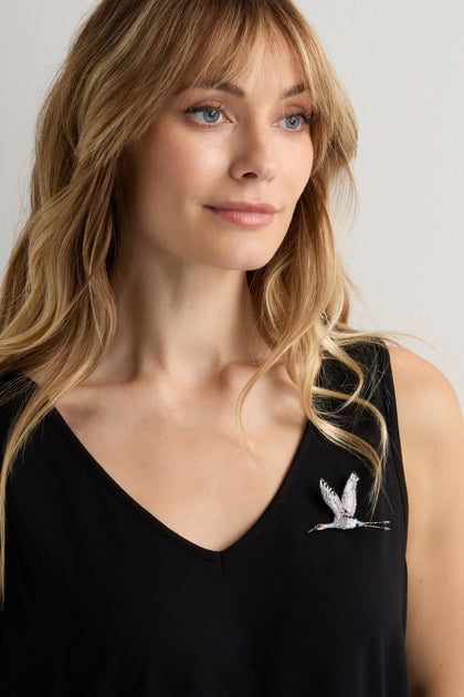 A woman with long blonde hair wears a black V-neck top styled with the Hand Beaded Sandhill Crane Brooch, featuring a nature-inspired flying bird design.