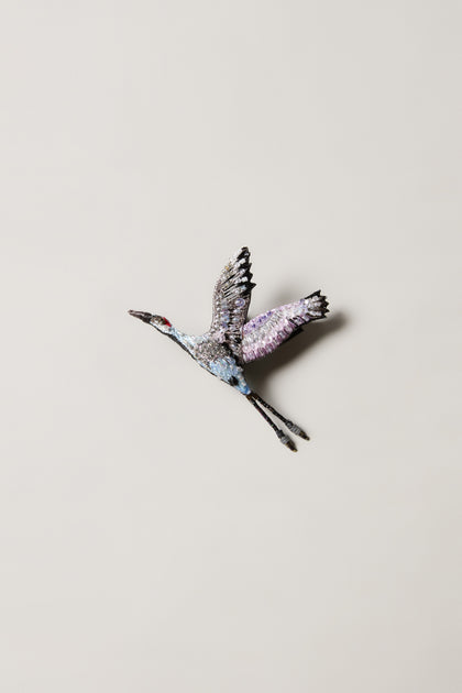 The Hand Beaded Sandhill Crane Brooch features outstretched wings in blue, purple, and black hues, displayed on a plain light background.