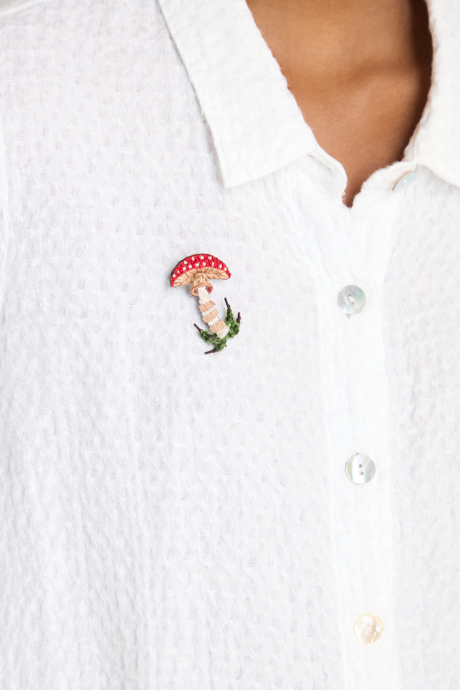 A person is wearing a textured white button-up shirt adorned with the Hand Embroidered Fly Mushroom Brooch and a grass pin on the left chest.