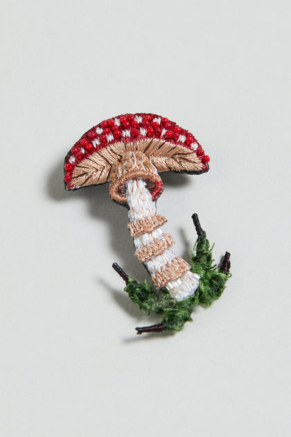 The Hand Embroidered Fly Mushroom Brooch is shaped like a red and white mushroom with a textured stem and green leaves at the base, shown against a light background.