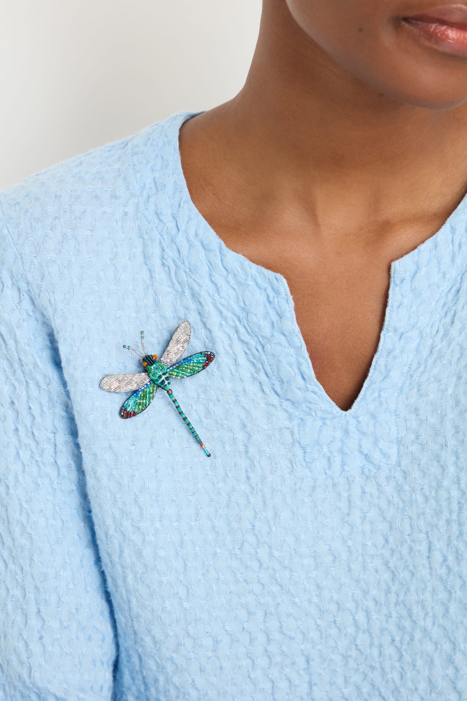 A person wearing a light blue textured top with the Hand Embroidered Oriental Greenwing Dragonfly Brooch pinned to the left side of their chest.