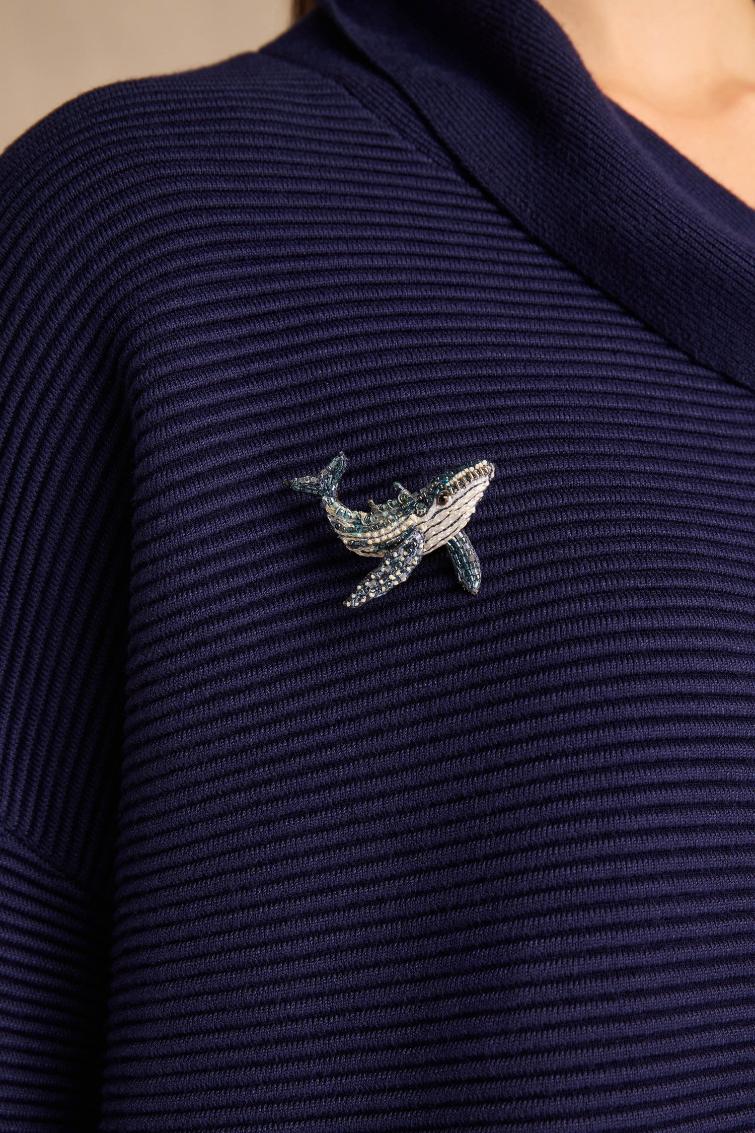 A Hand Beaded Humpback Whale Brooch is pinned to a navy blue, ribbed knit garment worn by a person, complementing the ensemble much like a Trovelore Brooch or an intricately hand-beaded dragonfly brooch would accentuate any outfit.