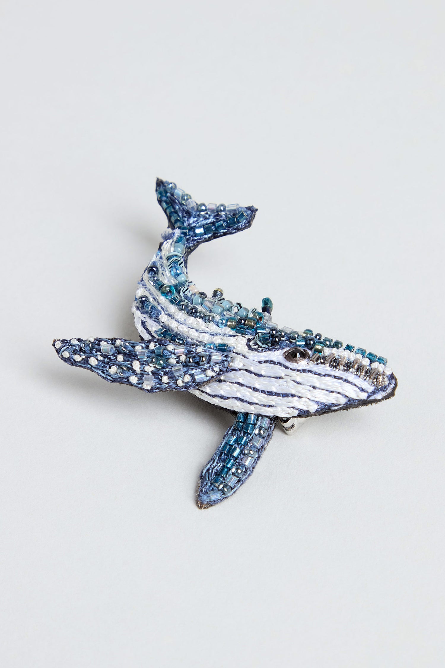 The Hand Beaded Humpback Whale Brooch, crafted from blue, white, and silver beads, is displayed on a light gray background.
