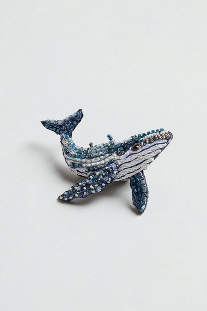 The Hand Beaded Humpback Whale Brooch features blue, white, and silver beads artfully arranged to resemble a whale, and is showcased against a simple light background—crafted in the unique style characteristic of Trovelore brooches.