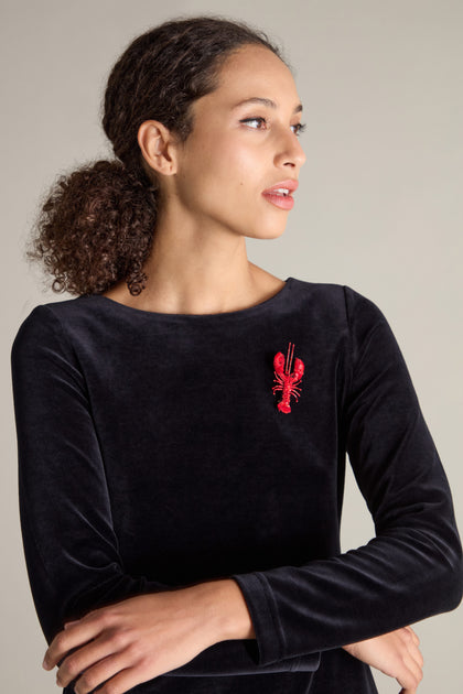 A person with curly hair wearing a black velvet top, adorned with a Hand Embroidered Lobster Brooch from Trovelore on the left side of the chest, featuring sequin and beaded embellishments.