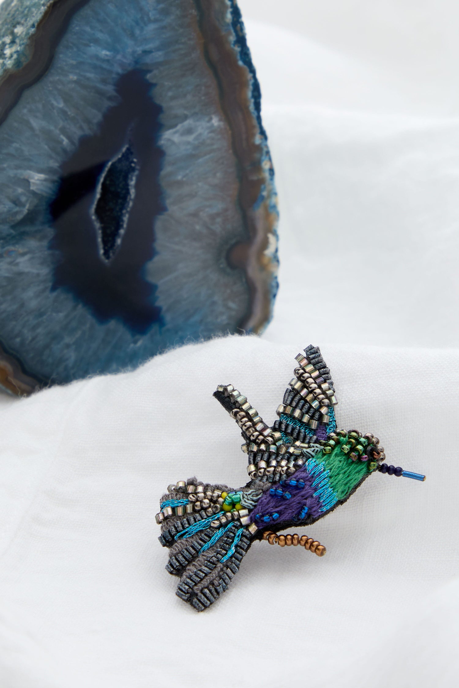 A Crowned Woodnymph Embroidered Brooch adorned with a colorful hummingbird, adding sparkle to the piece next to a beautiful agate.