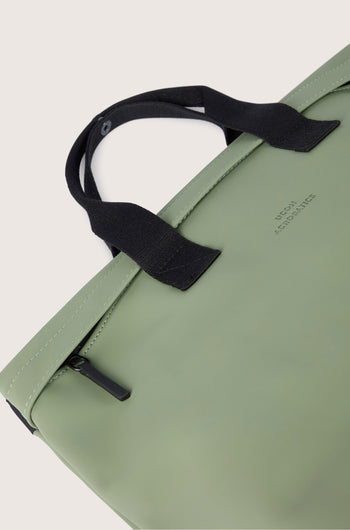 Close-up of a light green Eliza Roll Top Backpack with black handles and a visible zipper, showcasing its functional design. The words "JACQUEMUS" are subtly printed on the surface, highlighting its eco-friendly appeal by being made from recycled PET bottles.