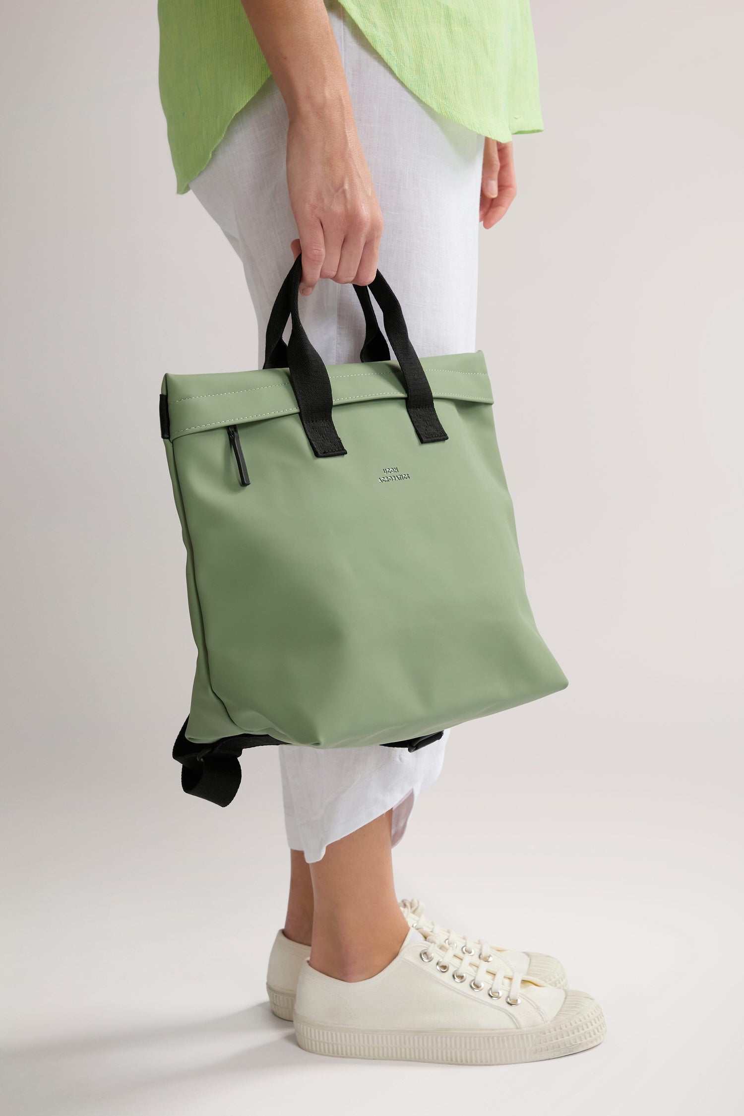 Person wearing a light green shirt and white pants holds an Eliza Roll Top Backpack made from recycled PET bottles with black handles and a zipper pocket, showcasing white sneakers.