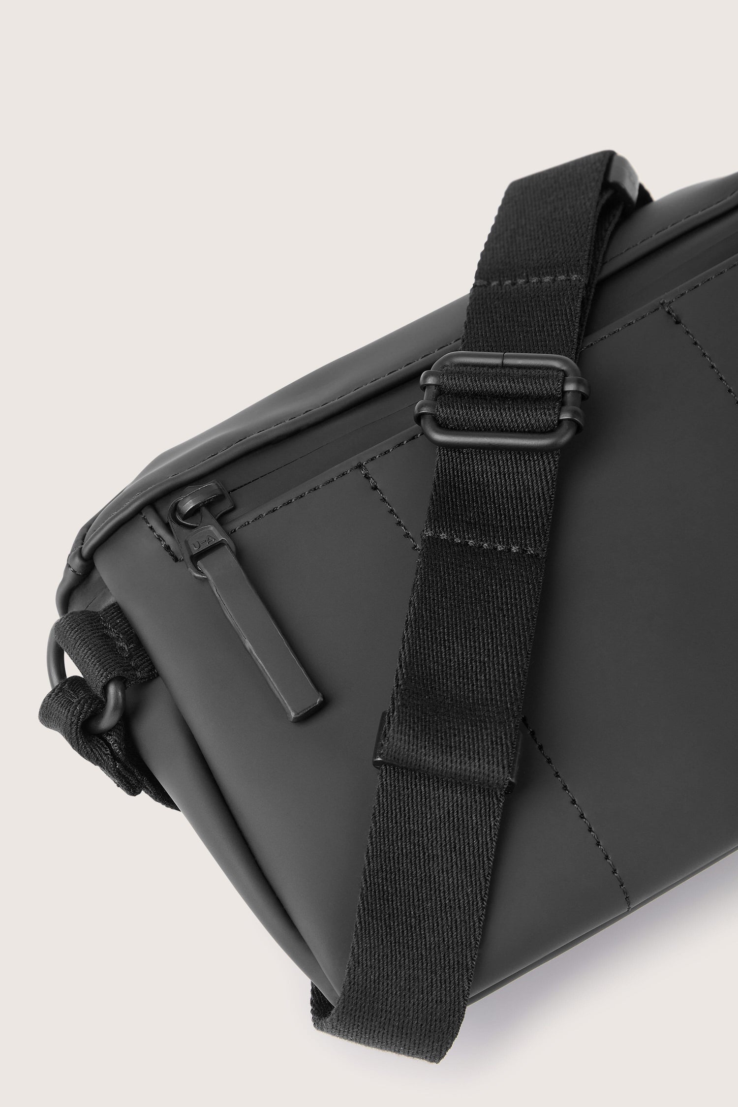 Close-up of the Jona Crossbody Bag, a black design with a quilted pattern and adjustable strap, crafted from water-repellent vegan tech material. The image highlights the zipper pull and strap buckle detail.