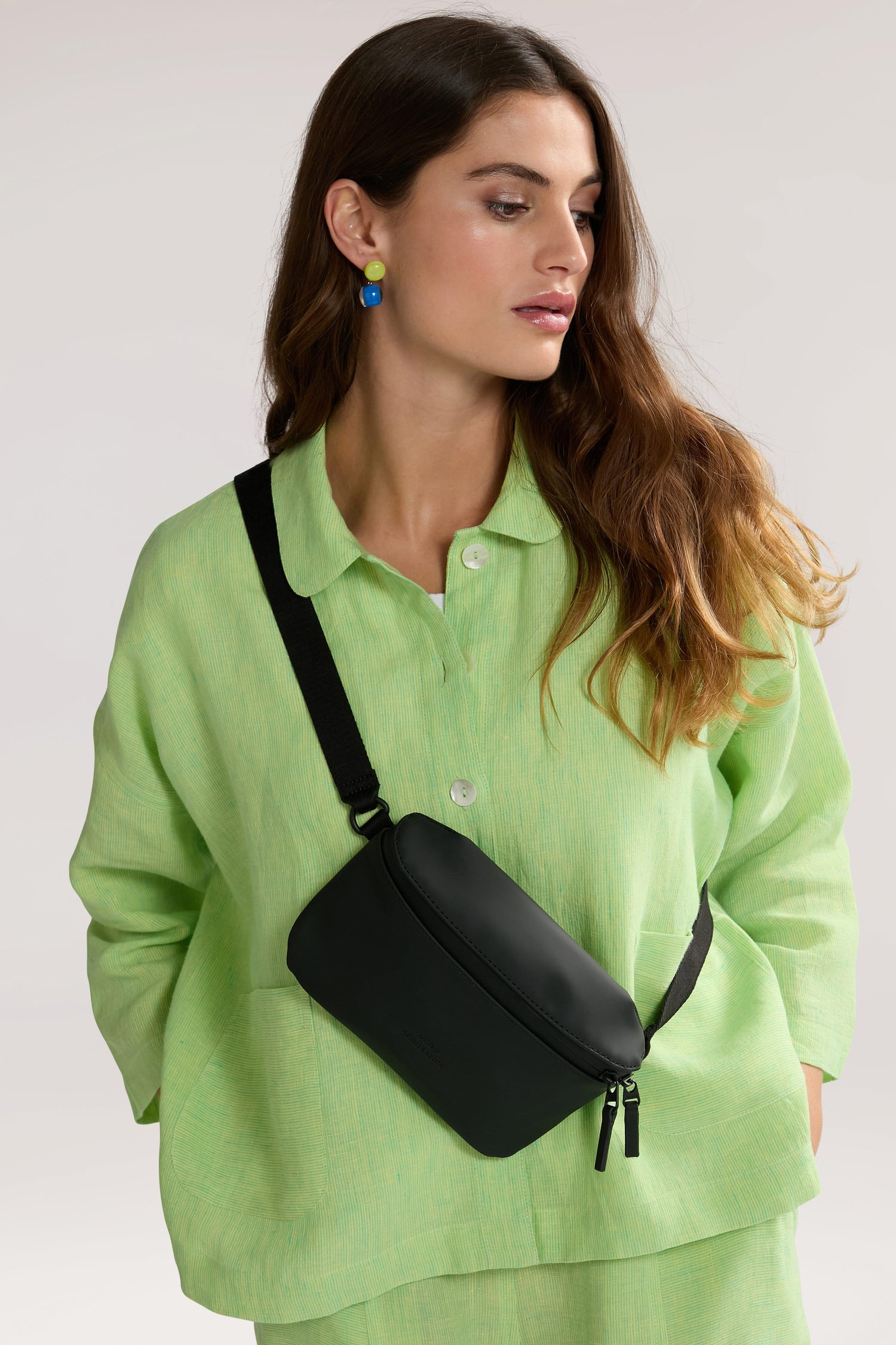 A woman with long brown hair wears a light green outfit and a water-repellent Jona Crossbody Bag. She has colorful earrings and is looking to her left against a plain background.