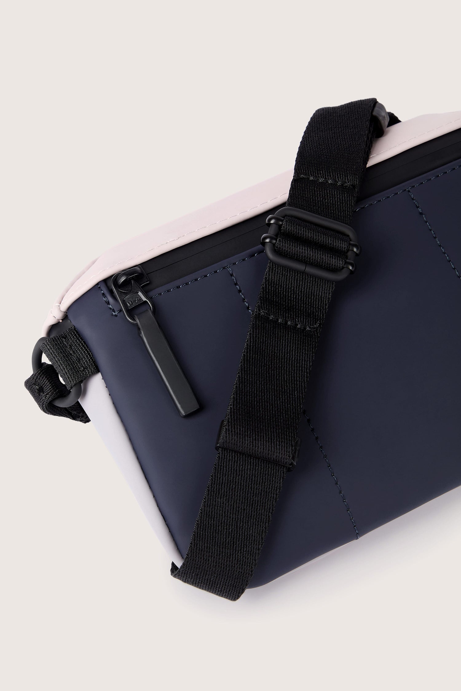 Close-up of the Jona Crossbody Bag in navy-blue with a black adjustable shoulder strap and a zipper. The bag, made from water-repellent, vegan tech material, features pale-pink accents along the edges.