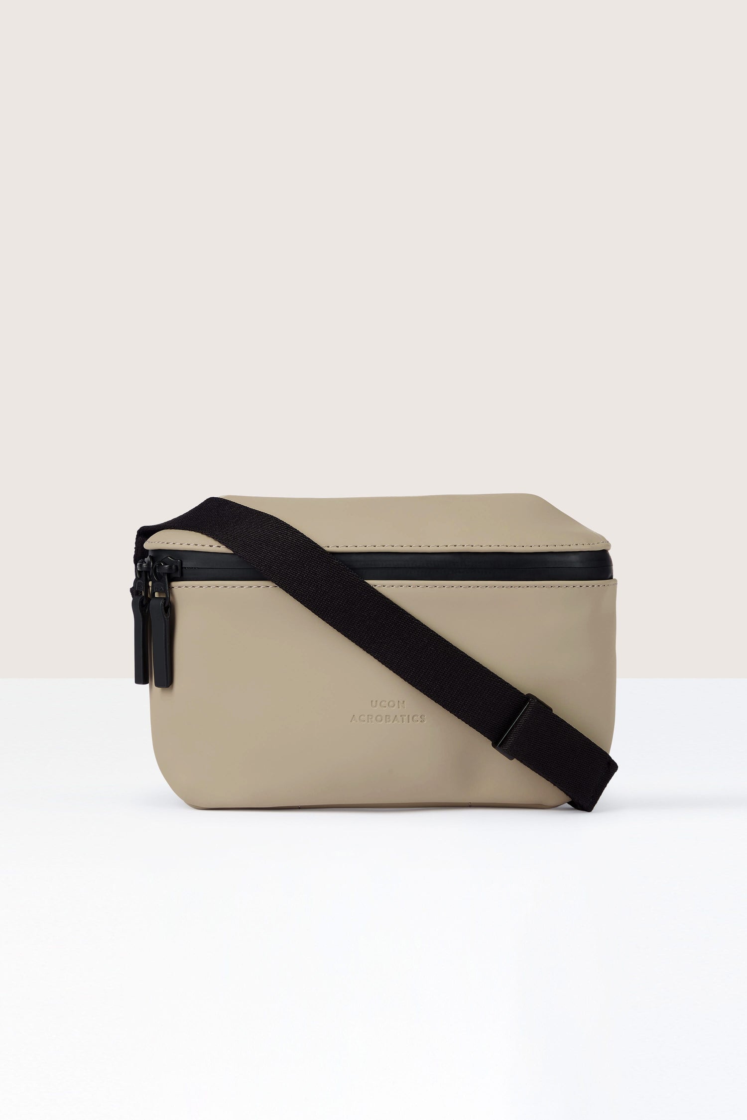 The Jona Crossbody Bag is a beige, rectangular crossbody bag crafted from vegan tech material, featuring black zippers and an adjustable black strap. It’s also water-repellent for added durability.