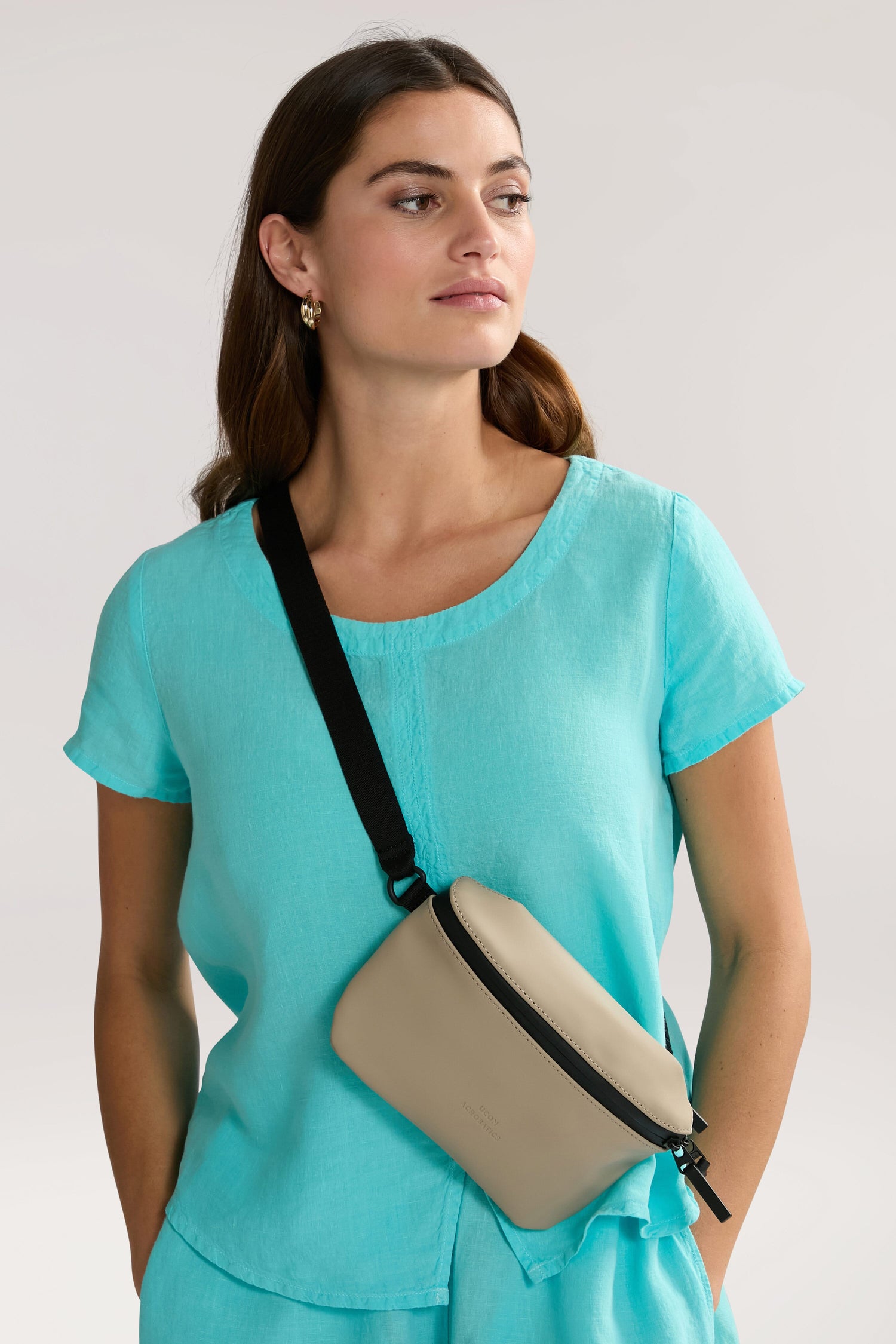 A woman in a light blue top with a beige Jona Crossbody Bag stands against a plain background, looking off to the side.