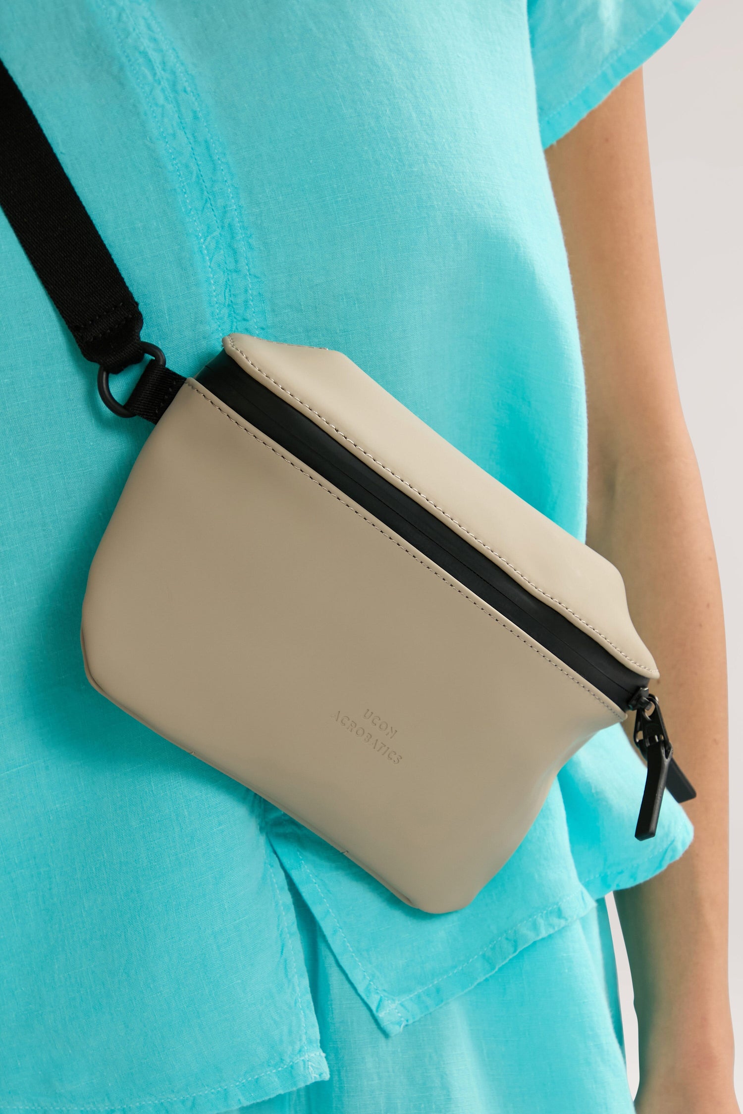 Person wearing a turquoise outfit with a beige Jona Crossbody Bag, featuring a black zipper and strap, made from vegan tech material.