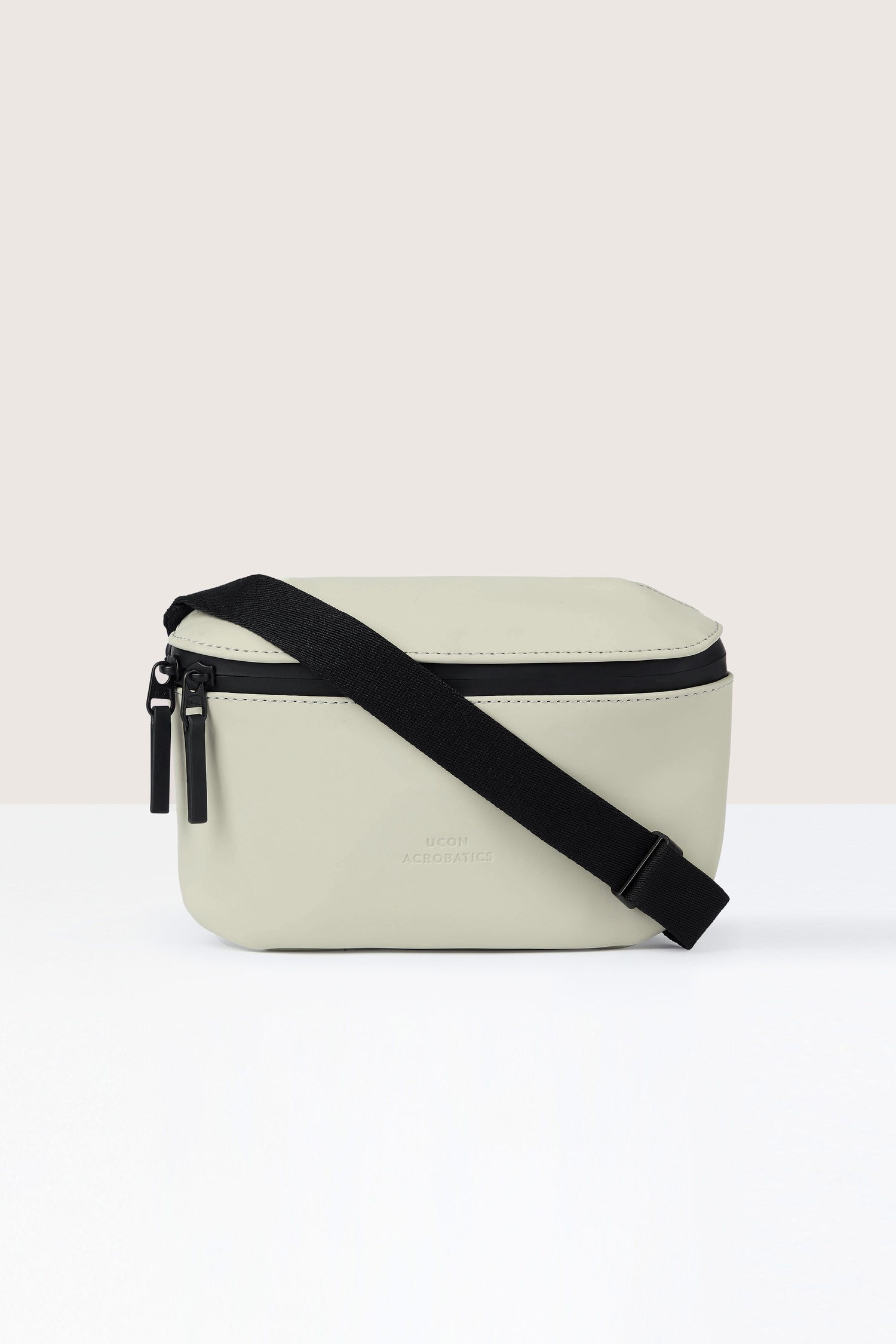 The Jona Crossbody Bag is a cream-colored, rectangular accessory made from vegan tech material. Featuring a black adjustable strap and two zipper pulls, it offers both style and function. Placed against a light background, its water-repellent surface ensures durability.