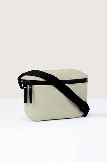 A light gray Jona Crossbody Bag with a black strap and double zipper closure, made from vegan tech material, positioned on a white surface against a neutral background.