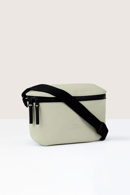 A light gray Jona Crossbody Bag with a black strap and double zipper closure, made from vegan tech material, positioned on a white surface against a neutral background.