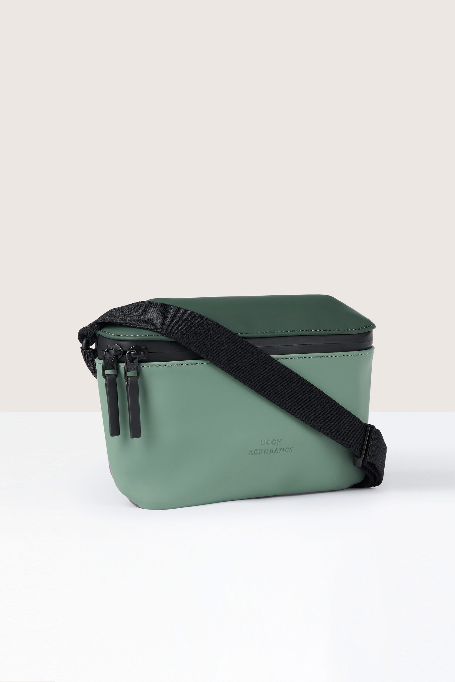 A small, green rectangular Jona Crossbody Bag made from water-repellent vegan tech material, featuring black zippers and a black adjustable shoulder strap on a white background.