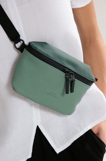 A person wearing a white top and black pants is seen with a green water-repellent Jona Crossbody Bag featuring two zippers.