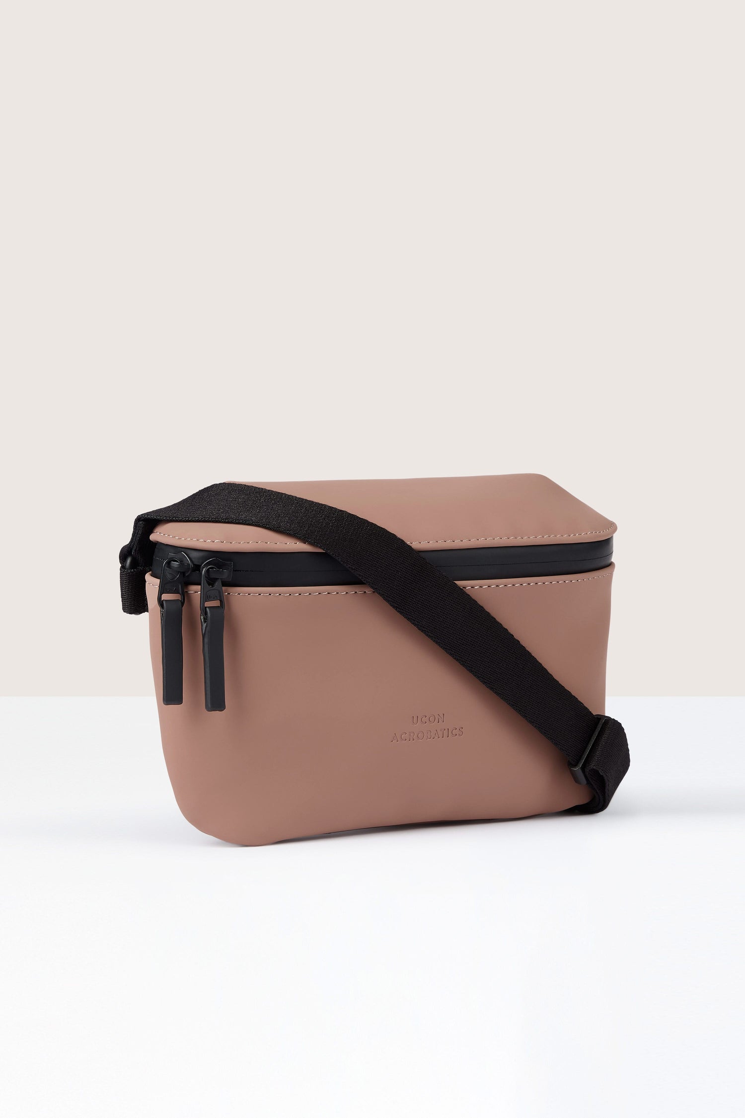 A pink rectangular Jona Crossbody Bag made from vegan tech material with black zippers and a black adjustable strap, positioned against a light-colored backdrop.