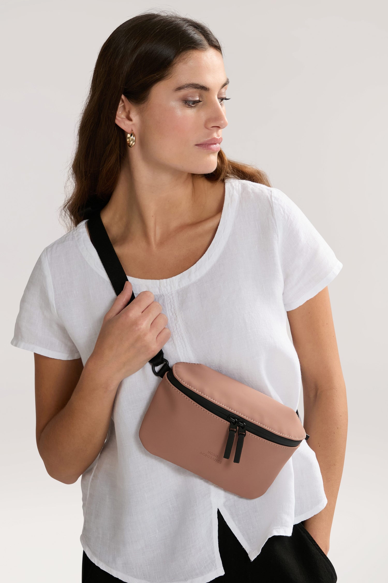 A woman in a white short-sleeved shirt and black pants holds a beige Jona Crossbody Bag with a black strap and zipper, crafted from water-repellent vegan tech material.