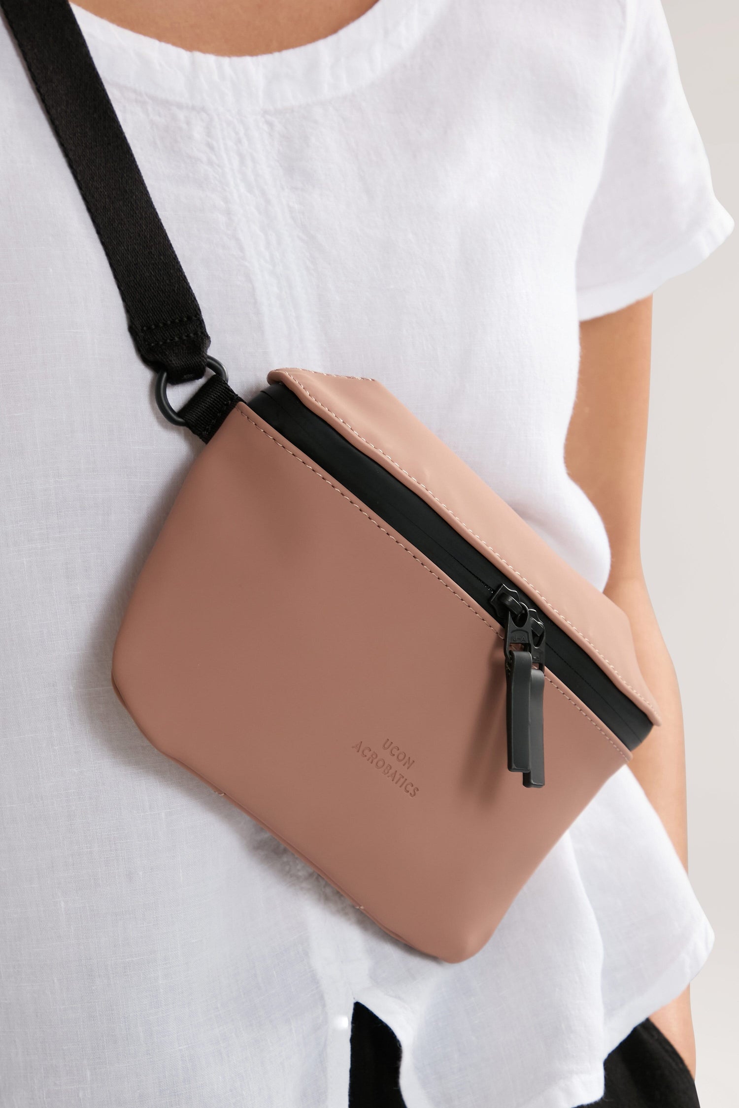 A person wearing a white top is seen with the Jona Crossbody Bag, made from vegan tech material, featuring a light brown design with a black strap and zipper.