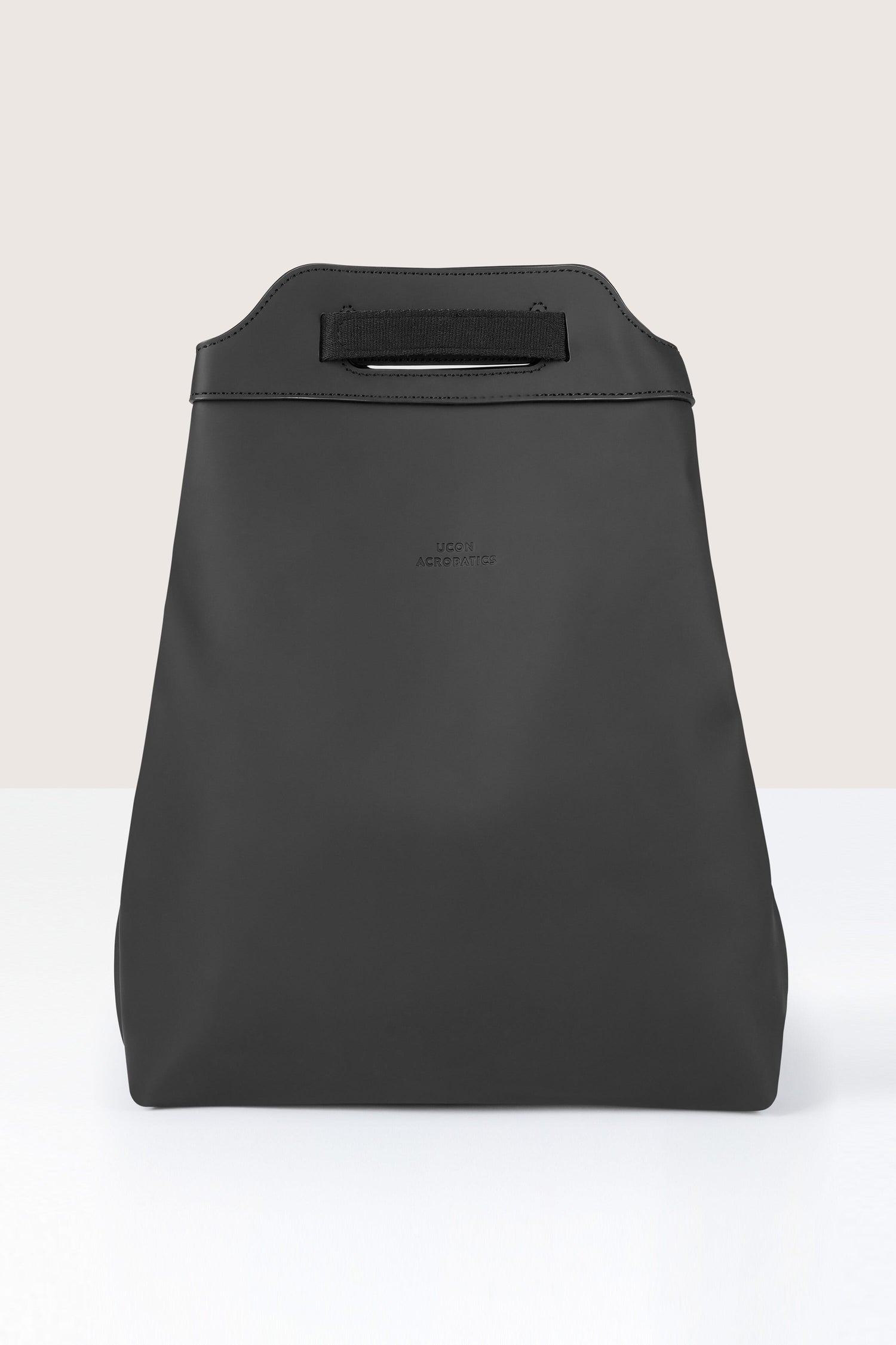 A black, minimalist Una Backpack made from recycled PET bottles with a built-in handle at the top, resting on a white surface against a beige background.