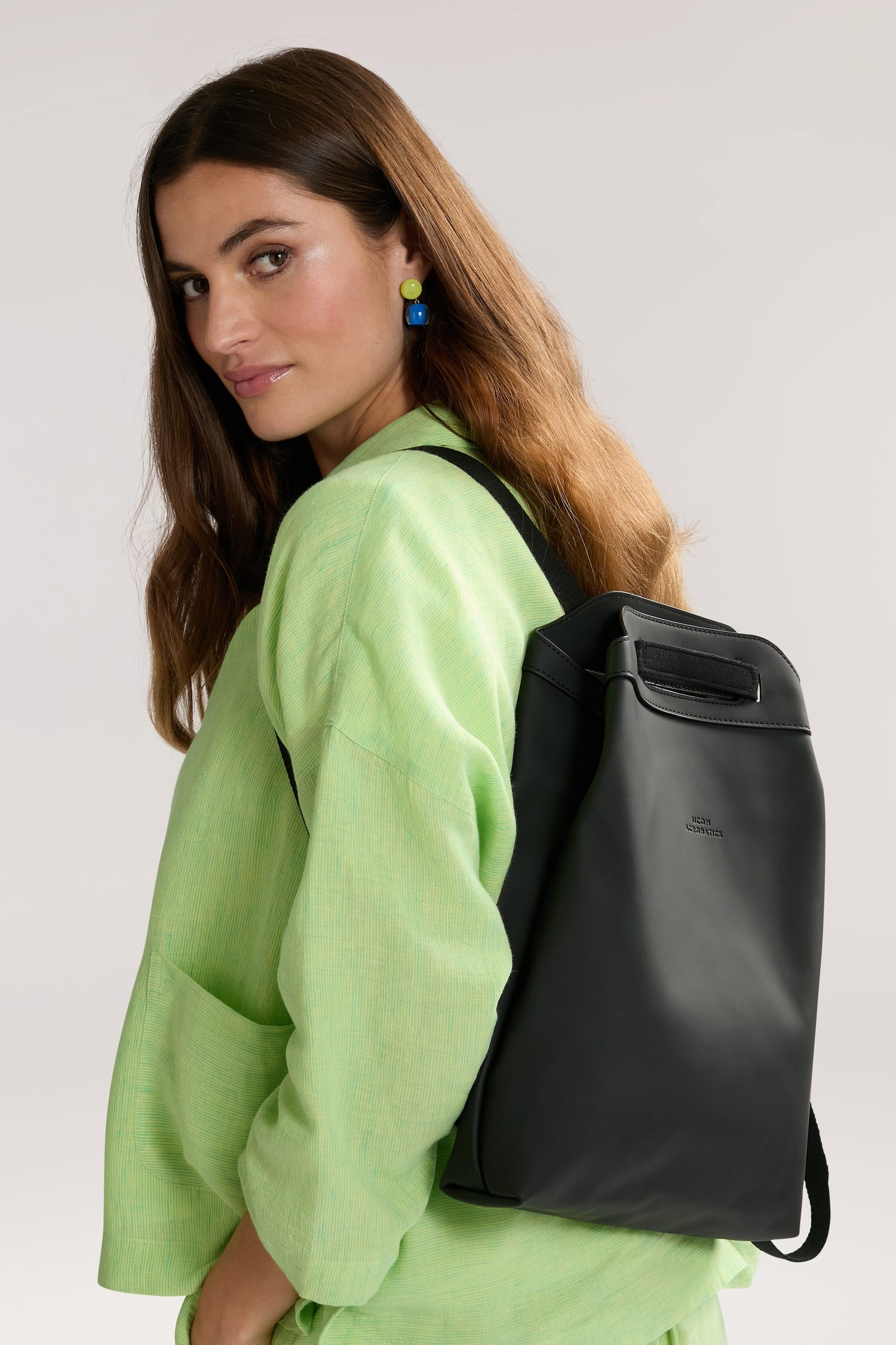 A woman with long brown hair, wearing a green outfit, glances over her shoulder while carrying the versatile Una Backpack, made from recycled PET bottles.