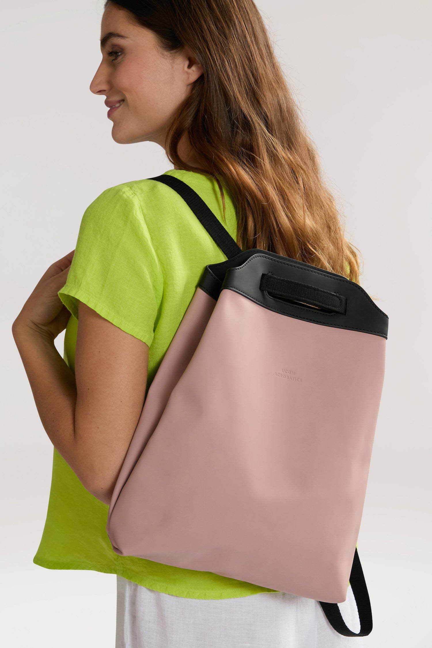 A person wearing a bright green top and white pants is carrying an Una Backpack, known for its versatile design, over their shoulder. The backpack, made from recycled PET bottles, stands out in pink and black against the plain, light-colored background.