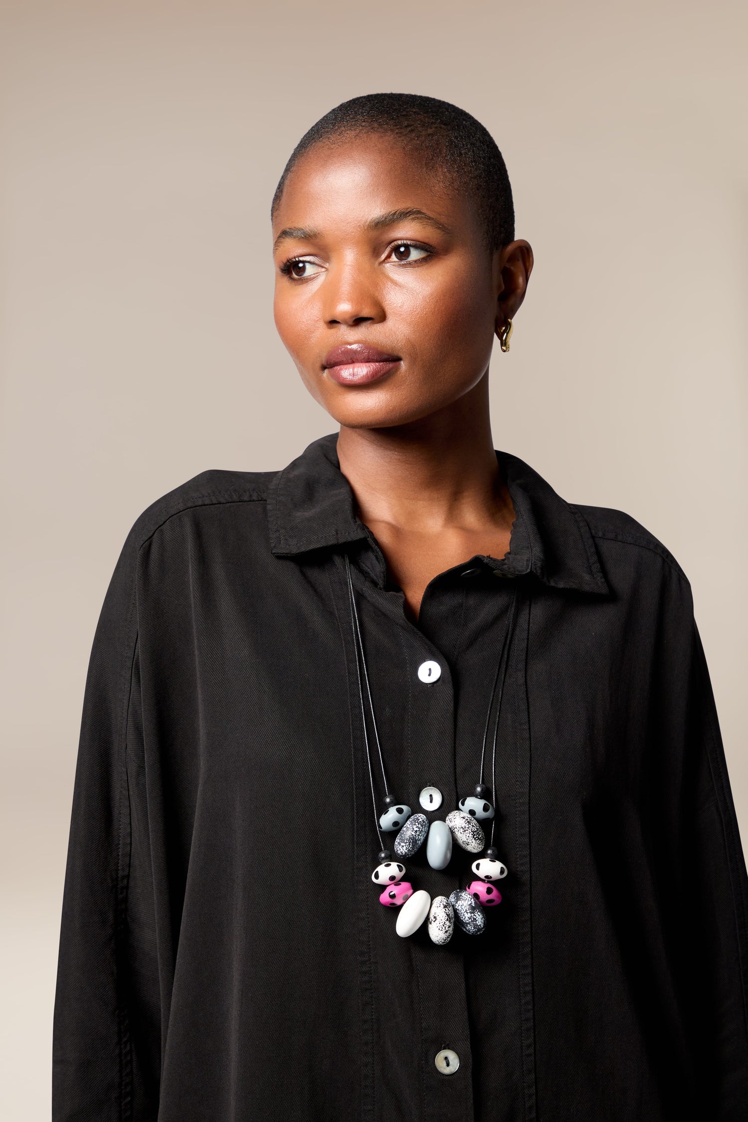 A person with short hair wears a black button-up shirt and the Dotty Two Strand Necklace, featuring large disc beads, while looking slightly to the side against a plain background.