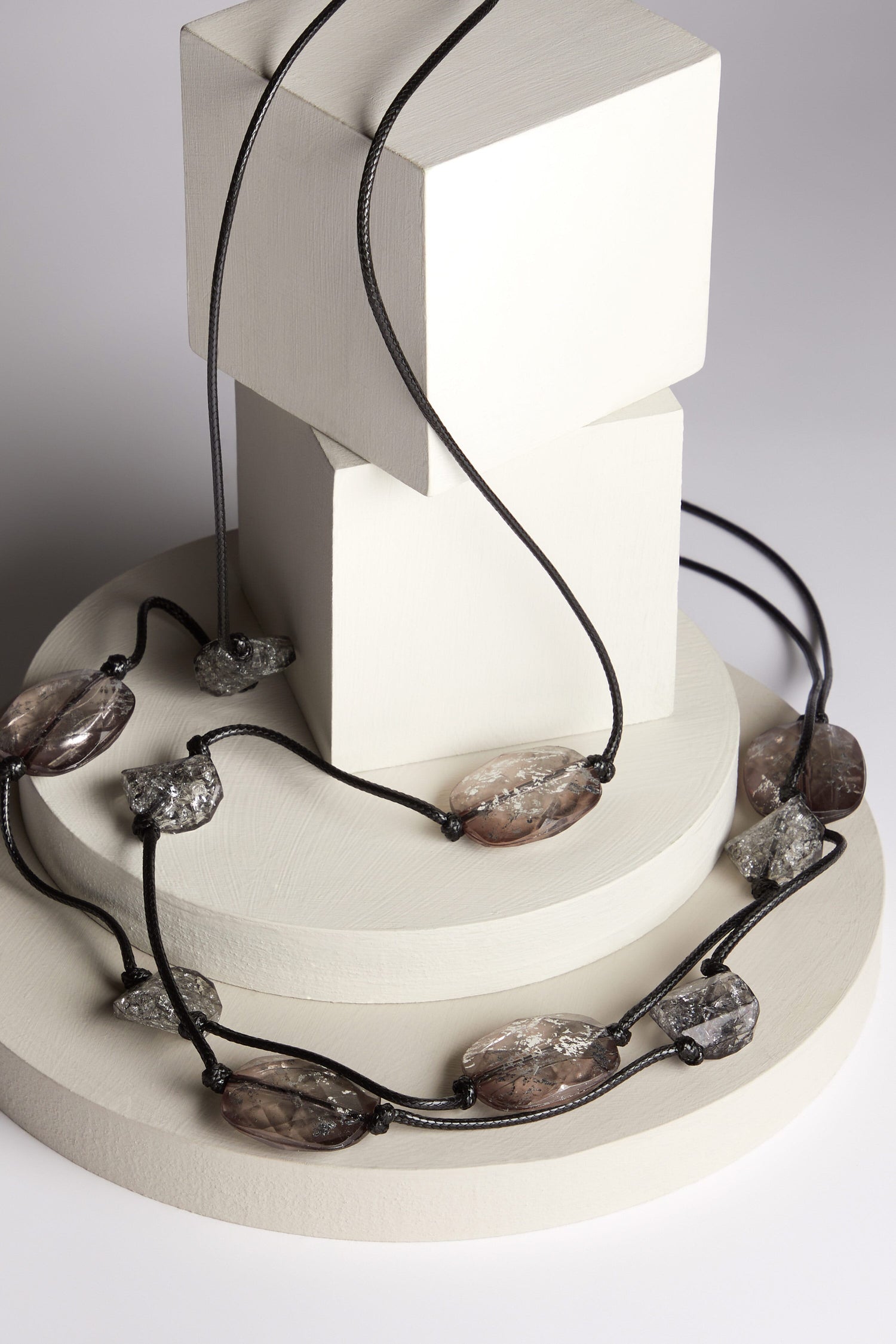 The Silver Leaf Pebble Necklace, the given product name, exhibits a dramatic flair, featuring translucent faceted beads in black and grey hues. It is elegantly showcased on top of a white pedestal.