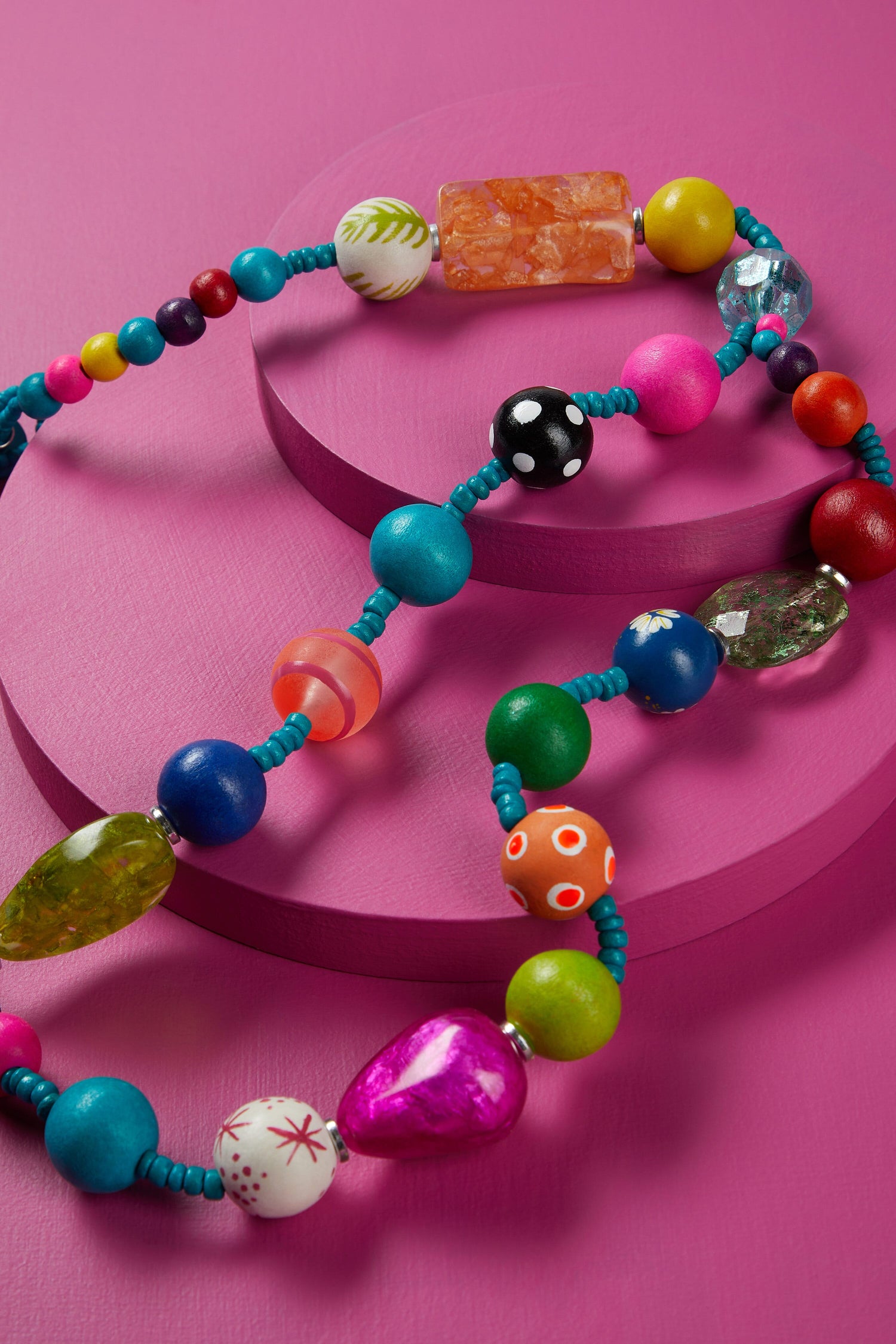 Colorful assorted whimsical beads on a Colour Pop Rainbow Spheres Necklace arranged in a spiral on a pink background.