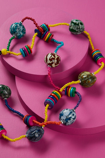 A Allsorts Rainbow Coco Bead Necklace with globe pendants arranged on pink surfaces with curving lines.