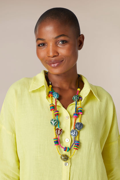 A smiling woman with a shaved head, wearing a yellow blouse and a multi-strand Allsorts Rainbow Coco Bead Necklace, looks directly at the camera.