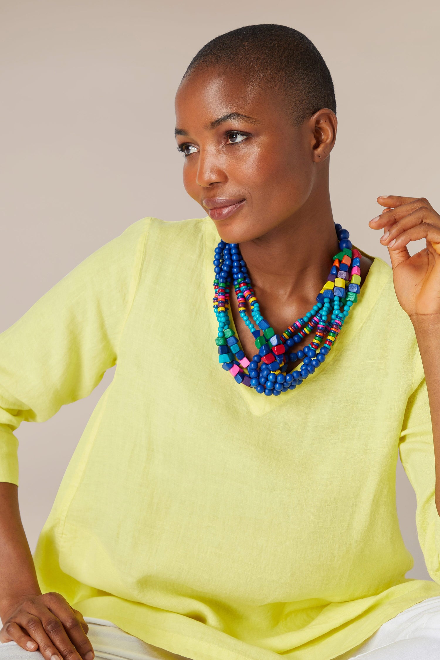 A woman with a shaved head wearing a yellow blouse and a Wooden Twist Bead Necklace made of saturated wooden round beads looks thoughtfully to the side.