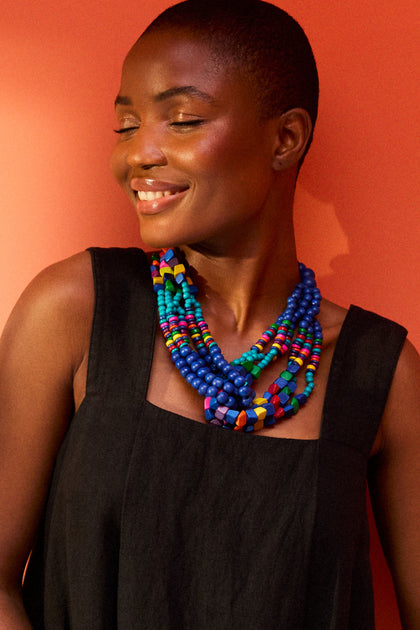 A smiling person with a shaved head wears a black sleeveless top and a Wooden Twist Bead Necklace against an orange background.