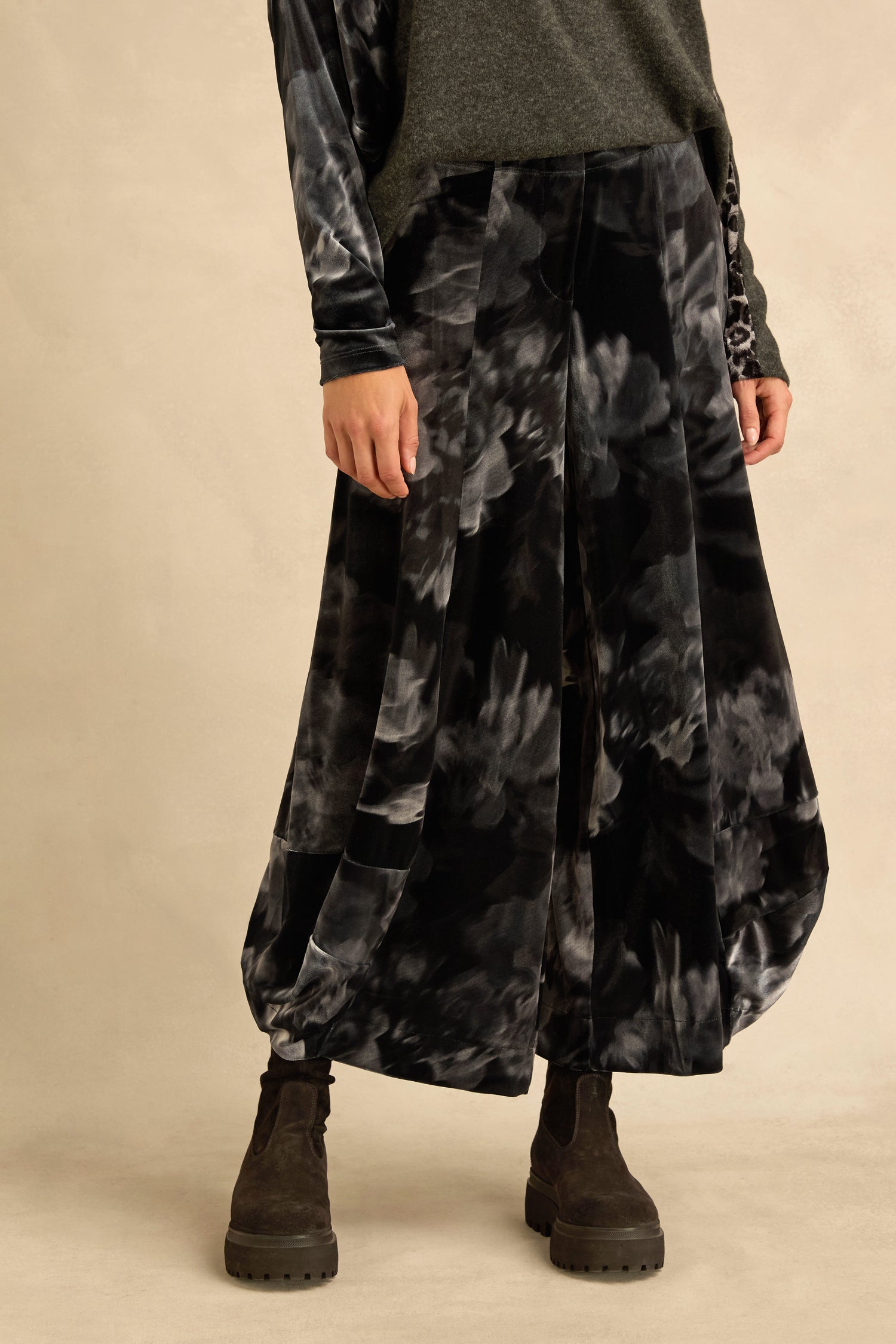 Person wearing a gray long-sleeve top, Blurred Floral Velvet Bubble Trousers, and black suede boots, standing against a beige background. Only the lower body is visible.