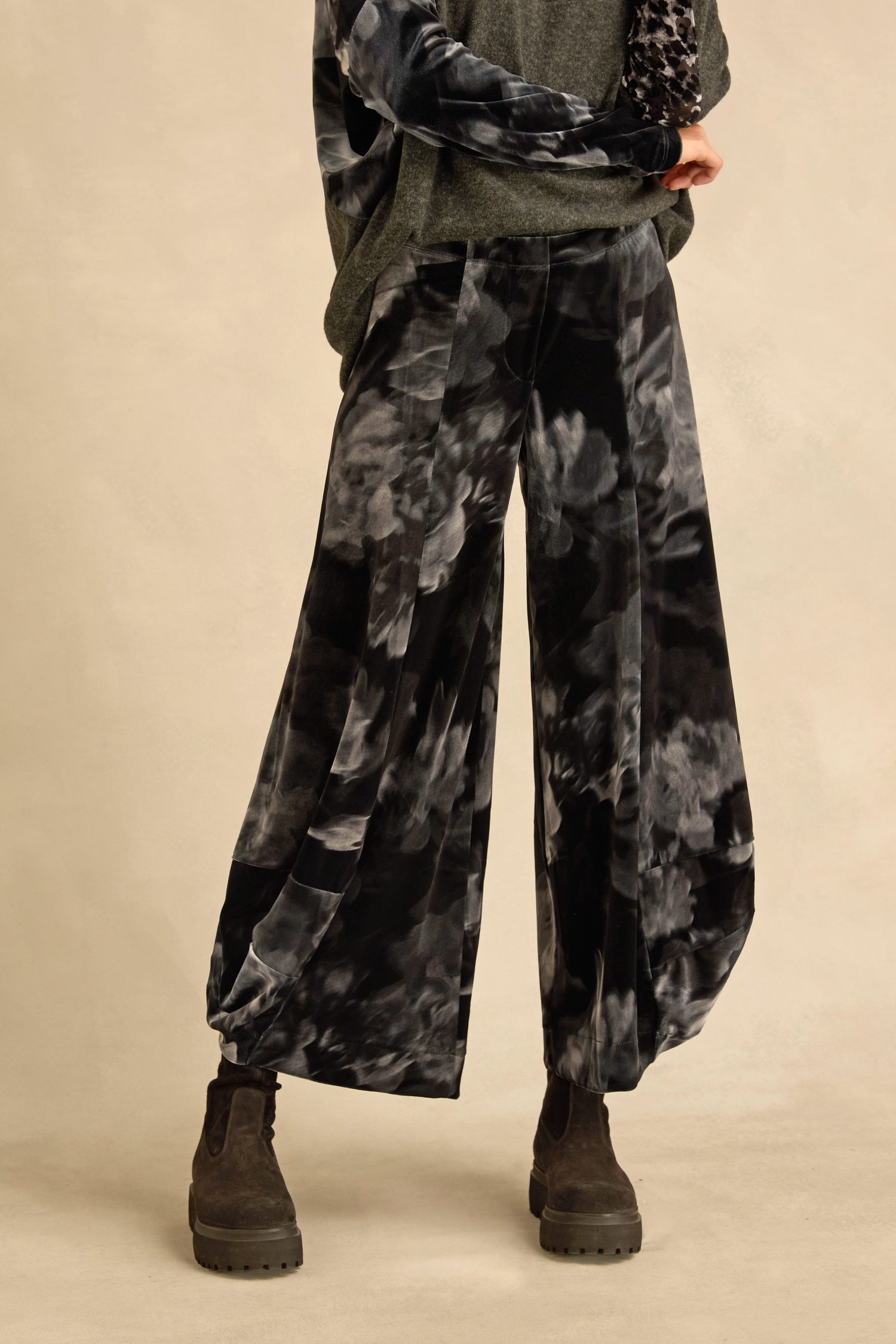 A person wearing the Blurred Floral Velvet Bubble Trousers—wide-leg pants featuring black and gray tie-dye with sculptural volume—paired with dark chunky boots and a gray long sleeve top, standing against a plain beige background.