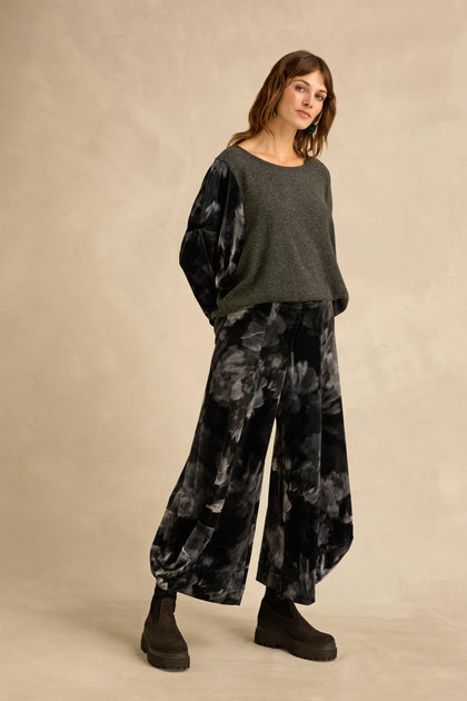 Woman standing against a beige background, wearing a loose grey sweater with sculptural volume, Blurred Floral Velvet Bubble Trousers, and black platform boots.