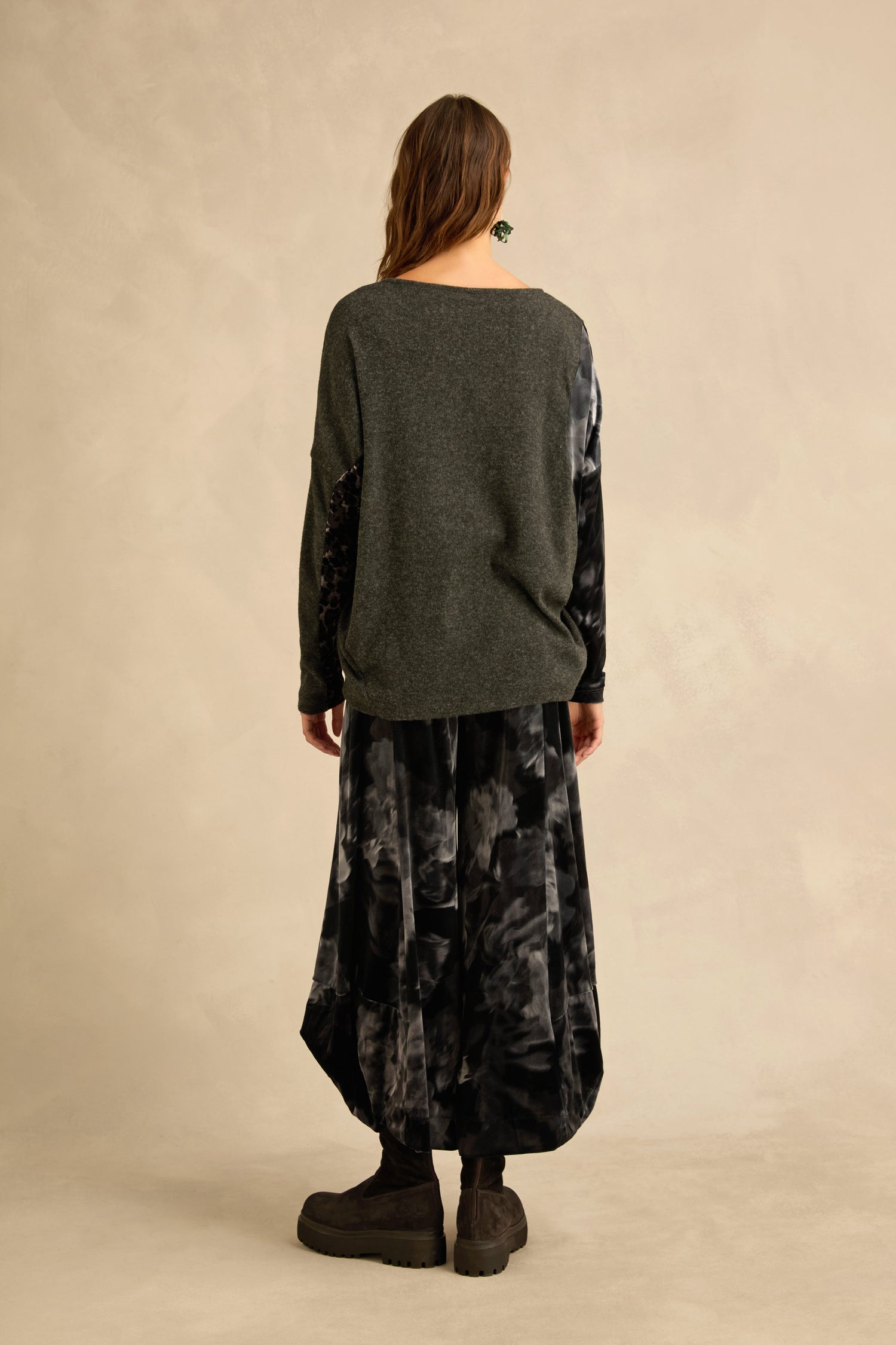 A woman stands with her back to the camera, wearing the Blurred Floral Jersey Top—a charcoal grey sweater featuring printed sleeves—paired with a black and gray patterned skirt and dark boots, set against a neutral background.