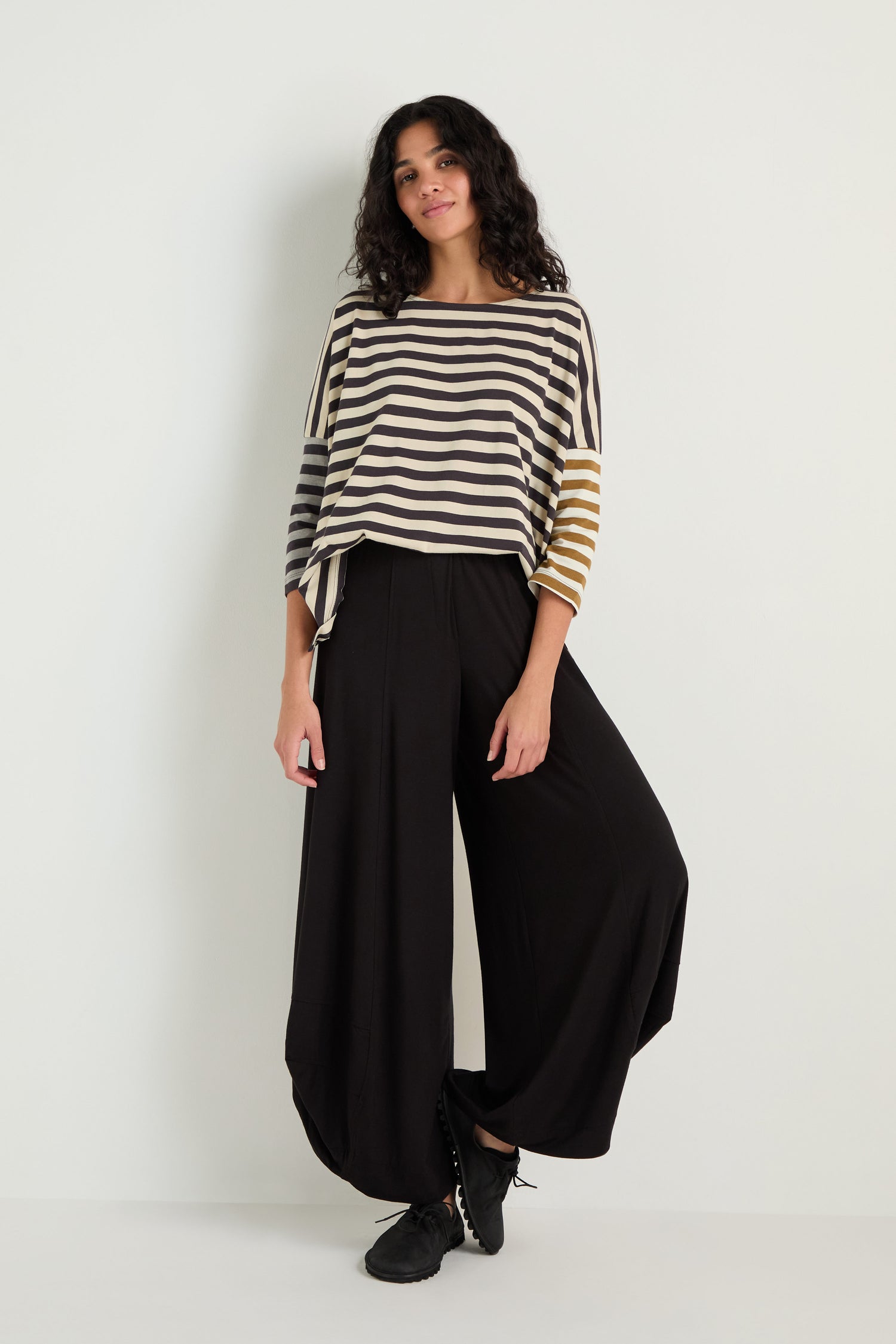 A person stands against a white wall wearing a loose striped top in black, white, and tan, paired with black Bubble Hem Trousers and black shoes.