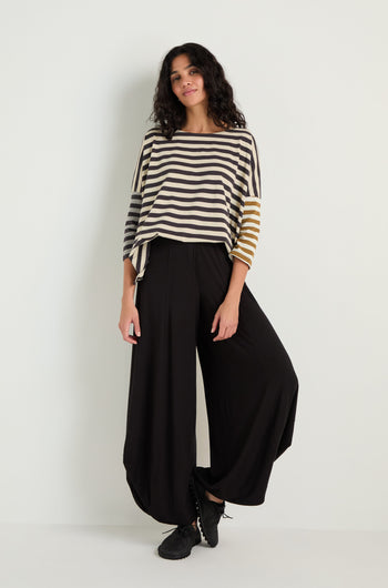 A person stands against a white wall wearing a loose striped top in black, white, and tan, paired with black Bubble Hem Trousers and black shoes.
